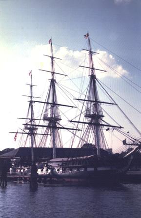 foreground: calm water; midground: docked three-masted ship at an angle; background: low building and sky